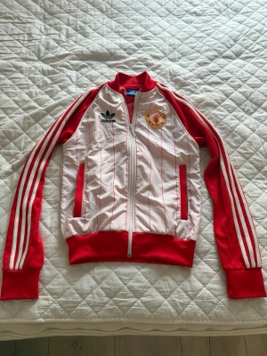 Manchester United Adidas track jacket - Snygg vit och röd track jacket från Adidas med Manchester United-logga på bröstet. Jackan har röda ränder längs ärmarna, röda muddar och dragkedja framtill. Tunn och sportig modell med diskreta röda ränder på kroppen. Perfekt för fotbollsfans! Jätte bra skick