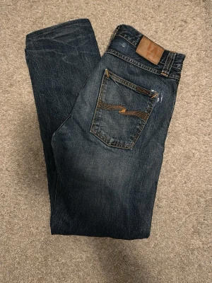 Mörkblå jeans från Nudie Jeans - Steddie edddie. Säljer ett par mörkblå jeans från Nudie Jeans med klassisk femficksmodell och snygga kontrastsömmar. Jeansen har en rak passform och detaljerad bakficka med Nudie-loggans brodyr. Perfekta för dig som gillar stilrena och tidlösa jeans.