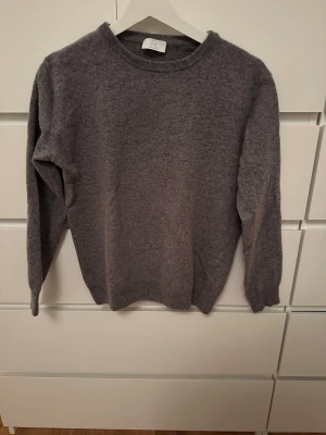 Grå stickad tröja Fillipi Roberto - Krymt så sitter som S. Mysig grå stickad tröja från   Fillipi Roberto med rund halsringning och långa ärmar. Tröjan har en enkel och stilren design, perfekt för lager på lager och passar till det mesta. Materialet ser ut att vara mjukt och värmande ullblandning.