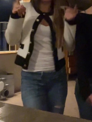 Vit kofta med svarta detaljer - Snygg vit kofta med svarta kanter och guldknappar. Kortare modell med rund halsringning och långa ärmar. Passar perfekt till jeans eller kjol för en trendig look. Materialet är mjukt och lite fluffigt