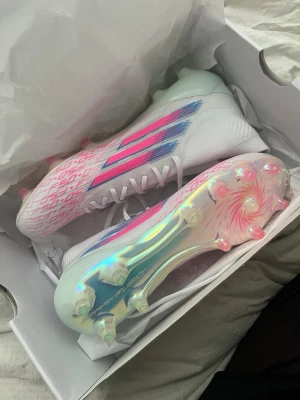 Adidas F50 Sparkfusion Elite FG/AG - Snygga Adidas F50 Sparkfusion Elite fotbollsskor i limited edition med vit bas, rosa och blå detaljer samt skimrande sula. Skorna har hög mid cut-design, snörning och FG/AG dobbar för spel på både natur- och konstgräs. Perfekta för dig som vill sticka ut på planen. Ordinarie pris 3200. Har endast prövat dom en gång.