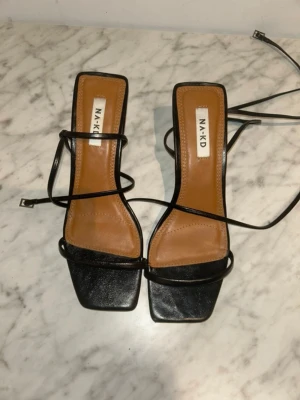 Svarta strappy pumps från NA-KD - Snygga svarta pumps från NA-KD med tunna remmar och fyrkantig tå. Skorna har en låg klack och långa snörningar som kan lindas runt ankeln. Bara använda en gång så dem är i jättebra skick🫶🏽