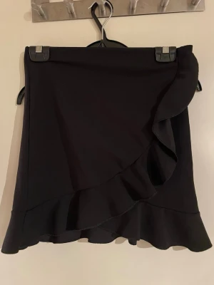 Svart omlottkjol med volang - Säljer en svart kortkjol med snygg omlott-design och volang längs nederkanten. Kjolen har en enkel och stilren look som passar perfekt till många olika outfits. Materialet känns mjukt och följsamt, och kjolen har en modern passform.