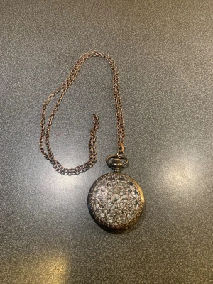 Vintage silverfärgad klocka med kedja - Säljer ett vintageinspirerat fickur i silverfärg med detaljerad kedja. Uret har ett vackert, ornamenterat lock med blommönster och genombruten design på framsidan samt graverade detaljer på baksidan. Perfekt accessoar för dig som gillar retrostil. Enbart som dekoration, klockan fungerar inte.