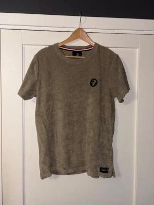 Beige frotté t-shirt från pace - Säljer en beige t-shirt från Pace i mjuk frotté. T-shirten har korta ärmar, rund halsringning och ett broderat märke med motiv på bröstet. Nedtill finns en liten Pace-label. Perfekt för dig som gillar unika material och stilrena detaljer.