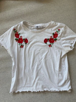 Vit t-shirt med broderade blommor - Vit t-shirt från FB Sister med broderade röda blommor på bröstet. T-shirten har korta ärmar, rund halsringning och vågig kant nertill. Perfekt för dig som gillar detaljer och vill ha en unik look. I storlek S