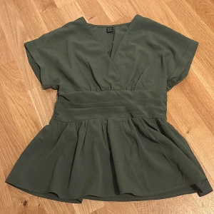 Olivgrön omlottblus med v-ringning - Säljer en olivgrön blus med v-ringning och omlottkänsla framtill. Toppen har korta ärmar, markerad midja med rynk och smock baktill samt ett lätt utsvängd nederdel. Perfekt för dig som gillar en feminin och avslappnad stil. Köpt från shein💚