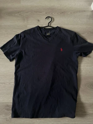 Mörkblå v-ringad t-shirt Polo Ralph Lauren - Klassisk mörkblå t-shirt från Polo Ralph Lauren med v-ringning och den ikoniska röda logon broderad på bröstet. Tillverkad i mjuk bomull och har korta ärmar för en avslappnad look.