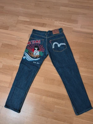 Evisu Mörkblå jeans med brodyr - Unika mörkblå jeans med färgglad brodyr på höger lår. Klassisk femficksmodell med raka ben och kontrastsömmar. Jeansen har måtten 96 cm i benlängd och 38 cm i midja. Perfekta för dig som vill sticka ut med coola detaljer och personlig stil.