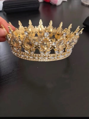 Guldig krona med strass och kristaller - Snygg guldig krona dekorerad med gnistrande strass och stora kristaller i olika former. Kronan har spetsiga detaljer och ett elegant, kungligt utseende som passar perfekt till maskerad eller cosplay. Tillverkad i metall med glittrande stenar för extra wow-effekt.