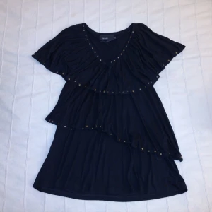 Oanvänd svart y2k topp/tunika med volanger och nitar - storlek M - Skitsnygg oanvänd svart topp/tunika med volanger och nitar. V-ringad och med fint fall. 100% viskos. Storlek M (40/42) men passar fler storlekar beroende på hur man vill att den ska sitta. Vet inte om det lossnat ett par nitar eller om de sitter lite ojämnt.