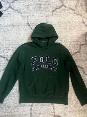 Grön hoodie från Polo Ralph Lauren - Mörkgrön hoodie från Polo Ralph Lauren med stor broderad POLO-logga och siffran 1967 på bröstet. Klassisk känguruficka framtill och mjukt material som känns skönt mot huden. Perfekt för en avslappnad och stilren look.