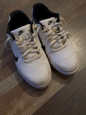 Vita Nike golf skor - Säljer ett par klassiska vita Nike Golfskor med svart swoosh på sidan. Skorna har lågt skaft, vita snören och svart insida. Perfekta för dig som gillar en clean och sportig stil. Materialet är syntet och textil.