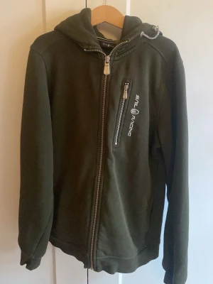 Grön hoodie från Sail Racing - Mörkgrön hoodie från Sail Racing med dragkedja framtill och en bröstficka med logga. Huva med dragsko och mjukt material som känns skönt mot huden. Perfekt för dig som gillar en sportig och avslappnad stil.