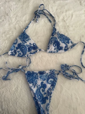 Blåvit bikini med paisleymönster - Snygg trekantsbikini i blått och vitt med paisleyinspirerat mönster. Både överdel och underdel har knytband för justerbar passform. Perfekt för stranden eller poolen!