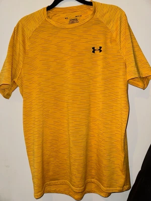 Gul tränings-tshirt från Under Armour - Sportig gul t-shirt från Under Armour med tunna svarta ränder och logga på bröstet. Tillverkad i ett lätt och snabbtorkande funktionsmaterial som passar perfekt till träning. Rund halsringning och korta ärmar för maximal rörelsefrihet.