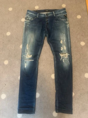 Replay jeans med slitningar blå - Säljer ett par blå skinny jeans från Replay med slitningar och hål på framsidan. Jeansen har knäppning med flera knappar och klassiska fem fickor. Materialet är denim och de har en tvättad look med ljusare partier. Perfekta för en avslappnad och trendig stil.
