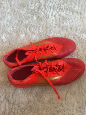 Röda F50 elite - Säljer ett par röda Adidas fotbollsskor med klassiska tre ränder i guld på sidan. Skorna har snörning och slitstark syntetisk ovandel samt fasta plastdobbar för spel på gräs. Perfekta för dig som vill sticka ut på planen och få bra grepp i spelet.