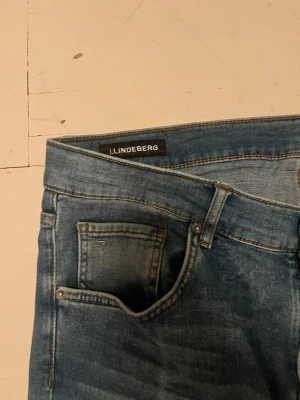 Blå slim fit jeans från J.Lindeberg - Snygga blå slim fit jeans från J.Lindeberg med klassisk femficksdesign och subtila slitningar. Jeansen har normal midja och raka ben, perfekta för en avslappnad men stilren look. Tillverkade i mjuk denim för skön komfort.