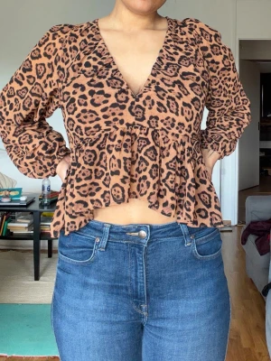 Leopardmönstrad blus med v-ringning - Säljer en snygg leopardmönstrad blus i brunt och svart med djup v-ringning fram och öppen rygg med knytband. Blusen har lång ärm och volang nedtill för en trendig look. Perfekt för dig som vill sticka ut lite extra!