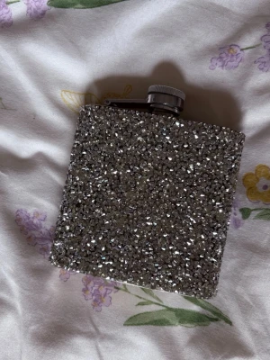 Silvrig fickplunta med glitter - Säljer en fyrkantig fickplunta täckt med glittrande silverfärgade stenar. Pluntan har ett skruvlock i metall och en kompakt design som gör den enkel att ta med. Perfekt accessoar för dig som gillar bling och vill sticka ut.