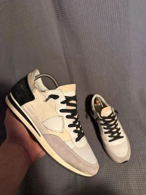 Philippe model  - Snygga sneakers från Philippe Model Paris i beige och svart med svarta skosnören. Ovandelen är i en mix av mocka och textil, med detaljer i vitt och grått. Klassisk låg modell med bekväm sula och ikonisk logotyp på sidan.