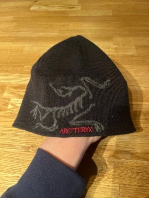 Svart mössa från Arc'teryx - Säljer en svart stickad mössa från Arc'teryx med deras ikoniska logga i grått och röd brodyr. Mössan är mjuk och smidig, perfekt för kalla dagar. Materialet är en skön ullblandning som håller värmen utan att bli för tjock.