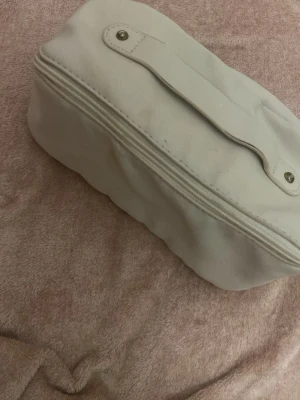 Vit necessär med handtag - En enkel och stilren vit necessär med dragkedja och praktiskt handtag på ovansidan. Väskan har en mjuk, rektangulär form och är tillverkad i ett slätt material, troligen syntet. Perfekt för att organisera småsaker eller smink i väskan. Den är smutsig i insidan som visas i bild