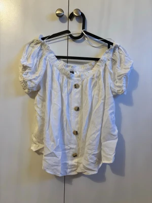 Vit offshoulder blus med knappar - Söt vit blus med offshoulder-design och puffiga korta ärmar. Blusen har flera dekorativa knappar framtill och en avslappnad passform. Perfekt för en trendig och luftig look.