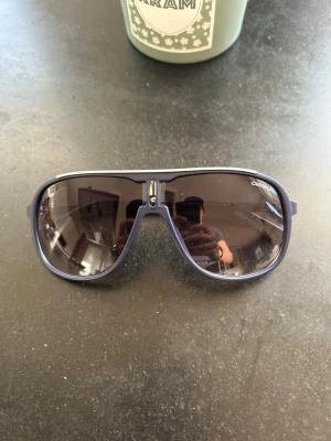 Blåa Carrera pilotbågar solglasögon - Snygga solglasögon från Carrera med blå pilotbågar och mörka tonade glas. Klassisk aviatorform med Carrera-logga på sidan. Ramen är i metall liknande material och glasen ger en cool look till alla outfits.
