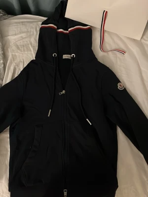 Marinblå hoodie från Moncler - Snygg marinblå hoodie från Moncler med dragkedja, huva och vita samt röda ränder på huvan och ärmsluten. Storlek S och har tyvärr ej kvitto men allt på bilderna ingår! Finns lite slitage som ej är synligt så skriv för fler bilder!
