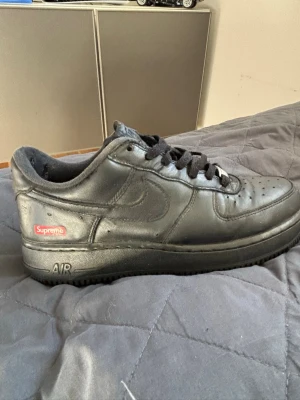 Nike Air Force 1 x Supreme svarta - Säljer ett par svarta Nike Air Force 1 i samarbete med Supreme. Skorna är låga och har en clean läderöverdel, svarta skosnören och Supreme-logga på sidan. Klassisk AF-1-detalj på snörningen och perforerad tå för extra ventilation.
