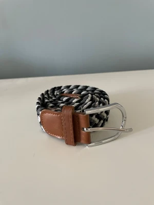Flätat bälte - Stretchigt grått, svart och vitt flätat bälte med bruna ändar och metallspänne. Cirka 100 cm långt (Se bild 4). Skriv gärna om du har nån fråga eller ett prisförslag.