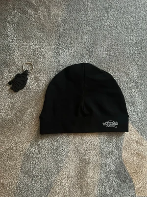 Svart mössa från syna world - Säljer en svart mössa i stretchigt tyg med en liten broderad logga i vitt framtill. Enkel och stilren design som passar till det mesta. Perfekt för dig som vill ha en clean look och gillar streetwear-vibbar.