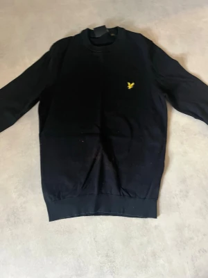 Svart sweatshirt från Lyle & Scott - Klassisk svart sweatshirt från Lyle & Scott med rund halsringning och den ikoniska gula fågel-loggan broderad på bröstet. Tröjan har ribbade muddar vid ärmslut och nederkant. Perfekt till ett avslappnat streetwear-utseende. Har en liten röd fläck på magen. Går säkert att få bort den, men jag har inte gjort det (fläcken drar såklart ner på priset)