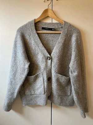 Grå kofta me knappar - Mysig grå kofta  med två stora fickor framtill. Koftan har v-ringning, 3 knappar och är tillverkad i ett mjukt, stickat material. Perfekt för lager på lager och enkel att matcha.