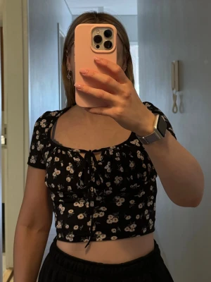 Blommig croppad topp med knyt - Säljer en svart croppad topp med vita blommor och knyt framtill.