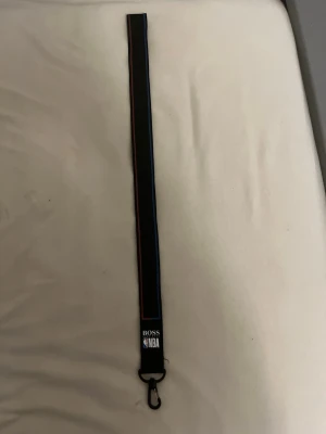 Svart nyckelband BOSS x NBA - Snyggt svart nyckelband från BOSS x NBA med röd och blå kant. Perfekt för att hålla koll på nycklar eller ID-kort. Metallhake i svart finish och tryckt logga från både BOSS och NBA. Tillverkad i slitstarkt tygmaterial. ”Jag har 3 stycken nya andra än de på bilden 