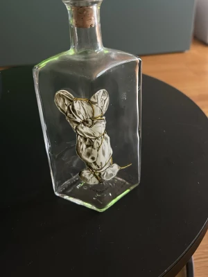 Dekorativ flaska med mus från Lasse Åberg  - Unik glasflaska med kork, innehållande en handgjord tygdocka insvept i vitt tyg och lindad med guldfärgad tråd. Flaskan är fyrkantig och transparent, perfekt som udda inredningsdetalj eller samlarobjekt. Nubbe flaska från Lasse Åberg 