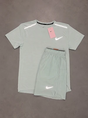 Nike Miller set - Säljer ett stilrent ljusgrått träningsset från Nike, bestående av en kortärmad t-shirt och matchande shorts. Båda plaggen har diskreta vita Nike-loggor och är tillverkade i ett lätt, snabbtorkande material som passar perfekt för träning.