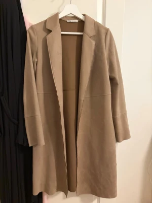Beige lång kappa från Zara - Stilren beige kappa från Zara med rak passform och klassisk krage. Kappan är lång och har en minimalistisk design utan synliga knappar eller fickor. Perfekt för dig som gillar en clean och modern look. Strl xs men passar även s och m