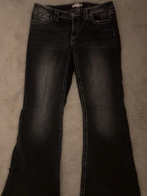 Svarta bootcut jeans med kontrastsömmar - Säljer ett par svarta bootcut jeans med tydliga vita kontrastsömmar och klassisk femficksdesign. Jeansen har en lätt tvättad look framtill och vida ben. Perfekta för dig som gillar retrostil och vill ha ett par jeans som sticker ut.