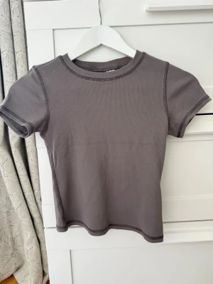 Grå ribbad kortärmad t-shirt - Snygg grå ribbad t-shirt med korta ärmar och rund hals. T-shirten har synliga sömmar och en tight passform som ger en modern look. Perfekt att matcha med jeans eller kjol för en enkel och stilren outfit.