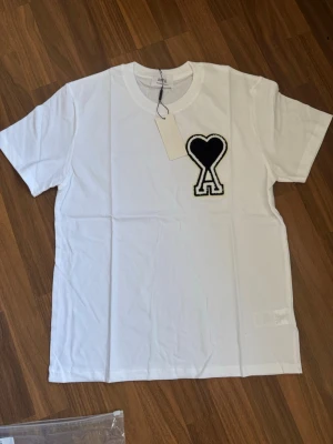 Vit t-shirt med svart hjärtdetalj Ami - Vit t-shirt från Ami Alexandre Mattiussi med en stor svart hjärtdetalj och bokstaven A på bröstet. Klassisk rund hals och korta ärmar. Tillverkad i mjuk bomull för en skön känsla och clean look.