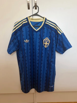Blå Sverige fotbollströja Adidas - Säljer en blå fotbollströja från Adidas med Sveriges landslagsemblem på bröstet. Tröjan har gula detaljer, tre gula ränder på axlarna och ett retroinspirerat mönster. Perfekt för match eller träning.