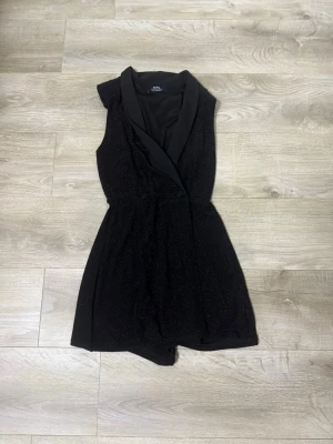 Svart glittrig ärmlös jumpsuit - Snygg svart jumpsuit med glittrigt tyg och bred krage i satinliknande material. Ärmlös modell med omlott framtill och korta ben. Perfekt för dig som vill sticka ut med en elegant och trendig look.