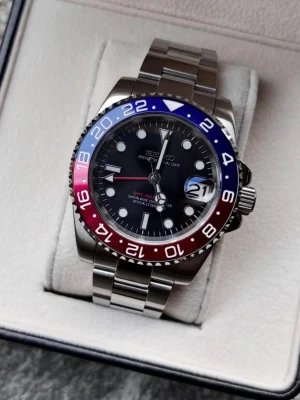 Seiko ‘Pepsi’ GMT- Master II (Oyster Bracelet) - -Movementζ: NH34A Seiko GMT -Case Size: 40mm -Water Resistance: 100m -Crystal: Sapphire Glass -Lume: Super Lume -Bracelet: Full link Jubilee & Oyster bracelet available (904L Stainless Steel) -Lifespan: 25+ years.                  Snygg Seiko GMT-klocka med rostfritt stålarmband och svart urtavla. Bezel i blått och rött ger en sportig look. Perfekt för dig som gillar klassisk men ändå färgstark stil.