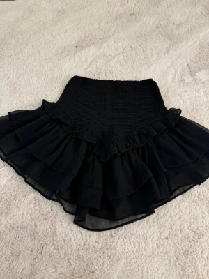 Svart volangkjol - Supersöt svart kjol från Gina Tricot med bred smock i midjan och flera lager volanger i tunn, luftig chiffong. Kjolen har en kort modell och ger en snygg siluett!