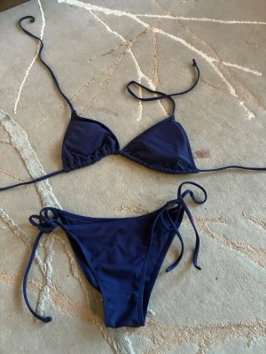 Mörkblå trekantsbikini med knyt - Snygg mörkblå bikini med trekantsöverdel och knyt i sidorna på både överdel och underdel. Klassisk och enkel design som passar perfekt till stranden eller poolen. Tillverkad i mjukt syntetmaterial för skön komfort.
