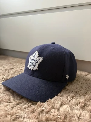 Mörkblå Toronto Maple Leafs keps - Snygg mörkblå keps med broderad Toronto Maple Leafs-logga framtill. Klassisk böjd skärm och justerbar passform. Tillverkad i ett mjukt tyg som känns bekvämt på huvudet. Perfekt för dig som gillar hockey och vill ha en stilren accessoar.
